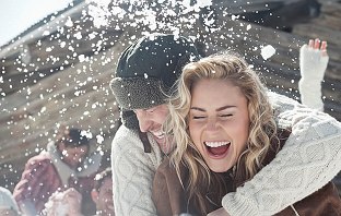 Junges Pärchen steht in einer winterlichen Region vor einer Hütte und genießt den günstigen Urlaub
