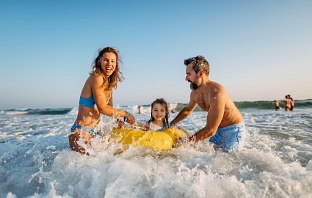 Junge Familie steht in im Meer und hält ihr kleines Kind auf einem gelben Schwimmreifen