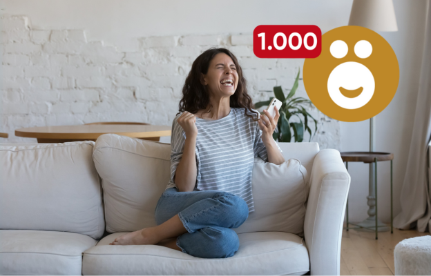 Frau sitzt auf der Couch und freut sich mit ihrem Handy in der Hand über 1.000 Ös 