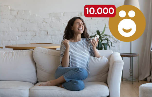 Frau sitzt auf der Couch und freut sich mit ihrem Handy in der Hand über 10.000 Ös 