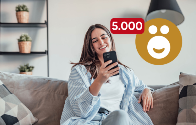 Frau sitzt auf der Couch und freut sich mit ihrem Handy in der Hand über 10.000 Ös 