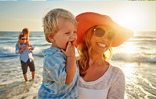 Frau steht mit ihrem Kind im Arm am Strand und genießt das schöne Wetter im günstigen Urlaub